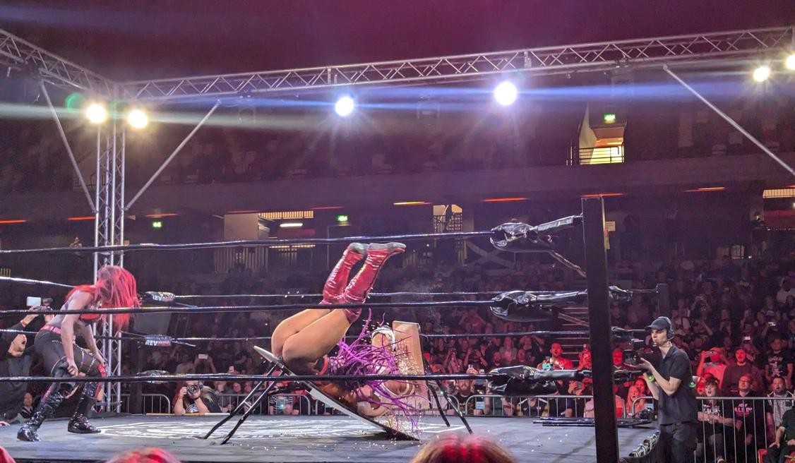 Alex Windsor puts Rhio through a thumbtack-covered table from the turnbuckle, as Mercedez Blaze looks on